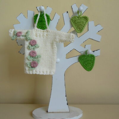 Hand knit minature jumper ornament in cream with pretty pink and green climbing rose applique hanging on a wooden tree photo prop.
