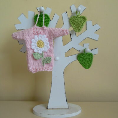 Hand knit minature jumper ornament in baby pink with a white daisy and green stem with leaves design hanging on a wooden tree photo prop.