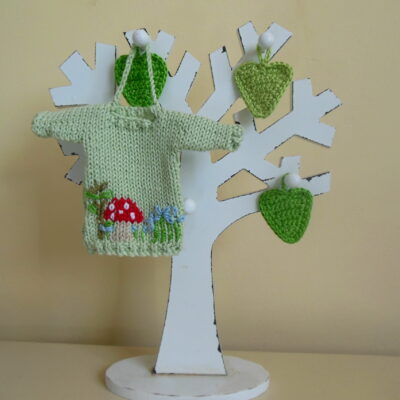 Hand knit minature jumper ornament in green with an embroidered red dotty mushroom and flowers scene hanging on a wooden tree photo prop.